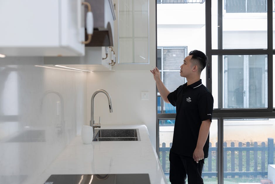 A man in black examines a kitchen cabinet inside a modern apartment with large windows.