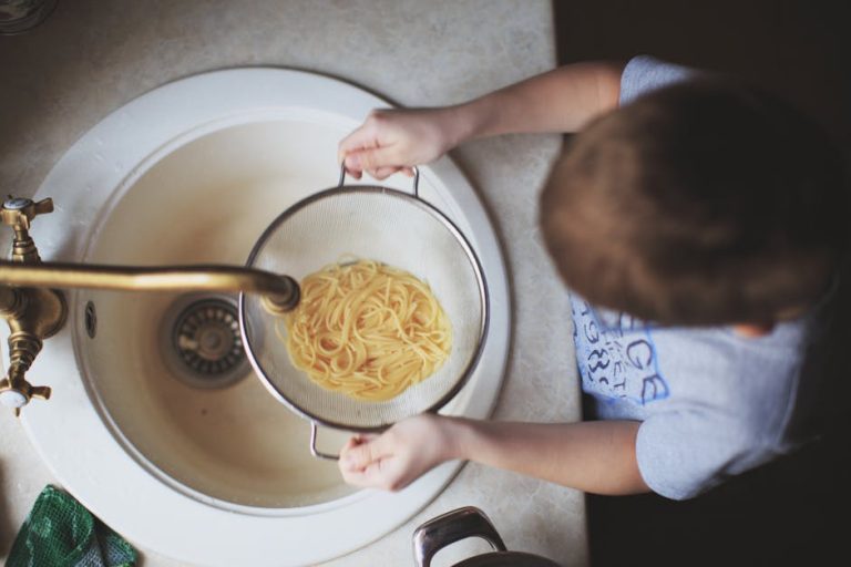 Overhead view of a child straining pasta in a kitchen sink.